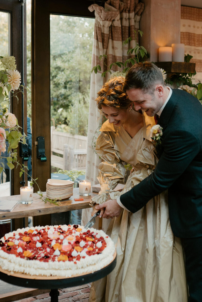 Bride and groom cake cutting moment with large Millefoglie Italian wedding cake decorated with fresh raspberries, edible flowers (shades of red and orange) and meringue kisses